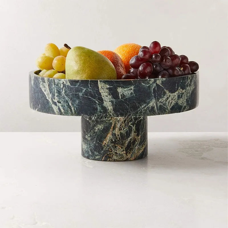 A round, premium fruit bowl with a stone finish holds a variety of fruits, including green and purple grapes, a pear, and oranges. The bowl has a green and white marbled pattern and is set on a white surface against a gray background.