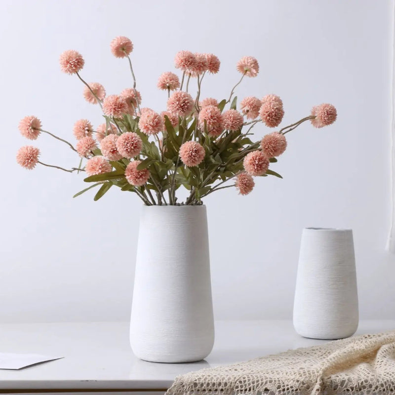 A bouquet of pink artificial flowers in a tall white vase sits next to a shorter white vase and a textured cloth.
