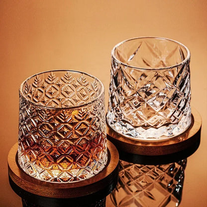 A pair of cut-crystal whiskey glasses, one filled with amber liquid and the other empty, sit on wooden coasters.