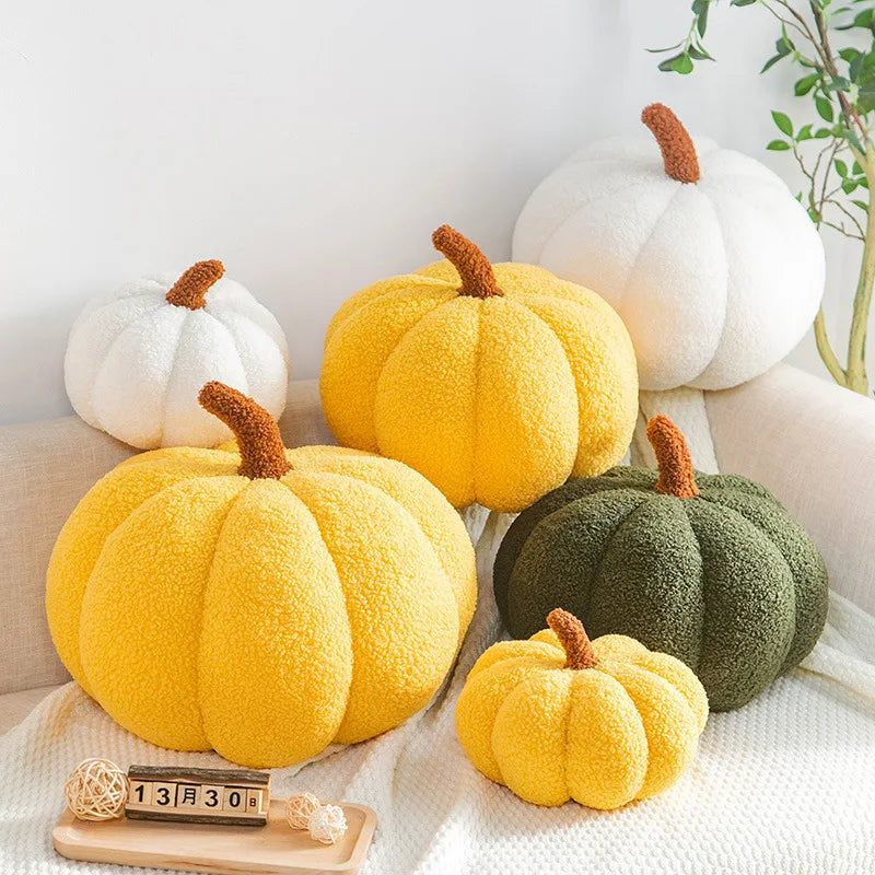 A cluster of plush, pumpkin-shaped pillows in shades of white, yellow, and green are arranged on a cream-colored sofa and draped with a white throw blanket.