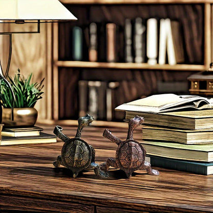 Two detailed turtle figurines, one brass and one bronze, on a wooden desk next to a stack of books and a lamp.