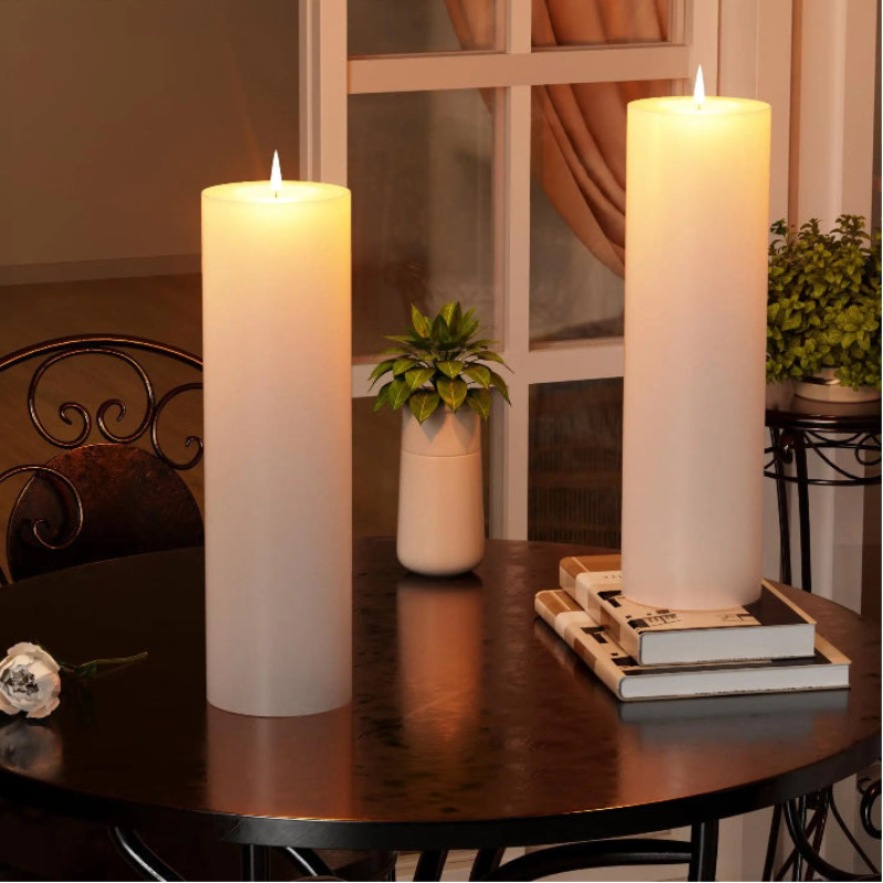 Two flameless pillar candles illuminate a dark room with warm light. A white rose and potted plant accent the candles.