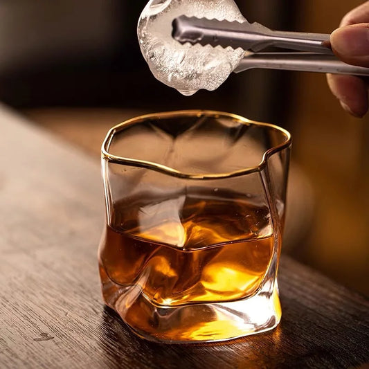 A pair of metal tongs holding a large, sculpted piece of ice over a unique, crumpled-looking glass filled with a brown liquid. The glass has a gold-rimmed edge.