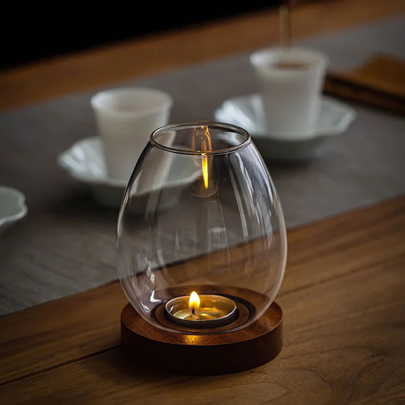 A warm and elegant glass candle holder with a wooden base, encasing a flickering tealight candle on a wooden table. In the softly blurred background, white teacups and saucers create a cozy and sophisticated ambiance.