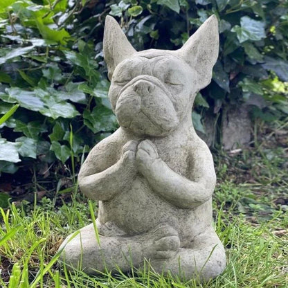 Gray stone French Bulldog statue meditating in a green garden.