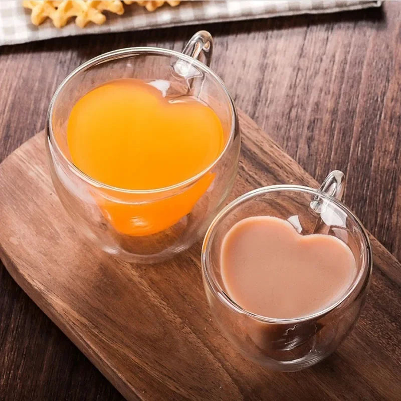 Two heart-shaped glass mugs on a wooden board; one with orange juice, the other with coffee.