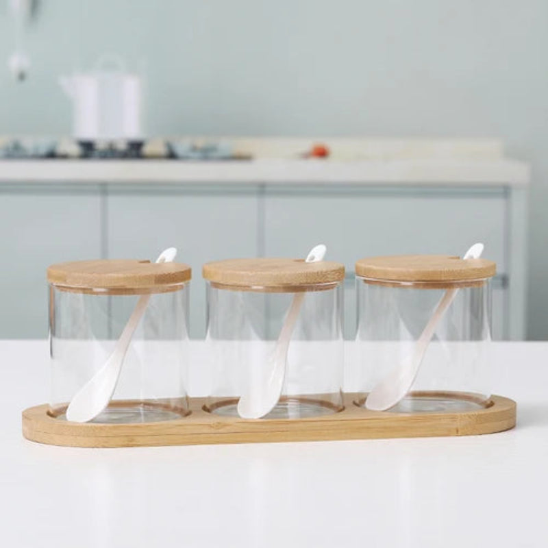 Three empty cylindrical glass jars with wooden lids and spoons, on a bamboo tray.