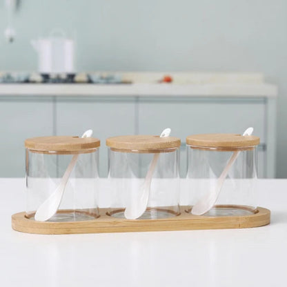 Three empty cylindrical glass jars with wooden lids and spoons, on a bamboo tray.