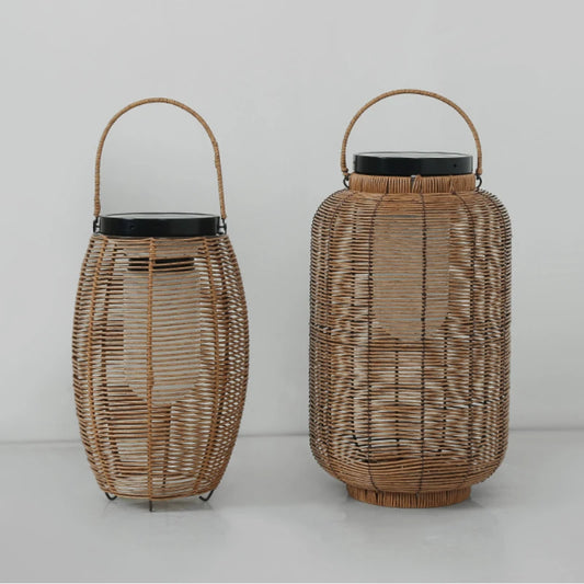 Two light brown, woven outdoor lanterns with dark tops and metal handles sit on a light surface against a plain background. The lantern on the left is taller and more oval, while the one on the right is shorter and more cylindrical with a slightly wider base.