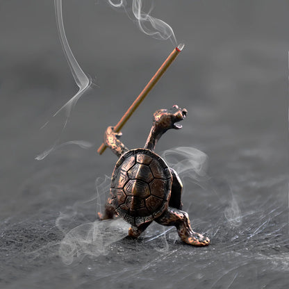 A close-up of a small, detailed bronze incense holder shaped like a turtle holding a burning incense stick like a sword, with smoke swirling around it.