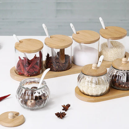 Seven assorted glass spice jars with wooden lids and spoons, on bamboo trays.