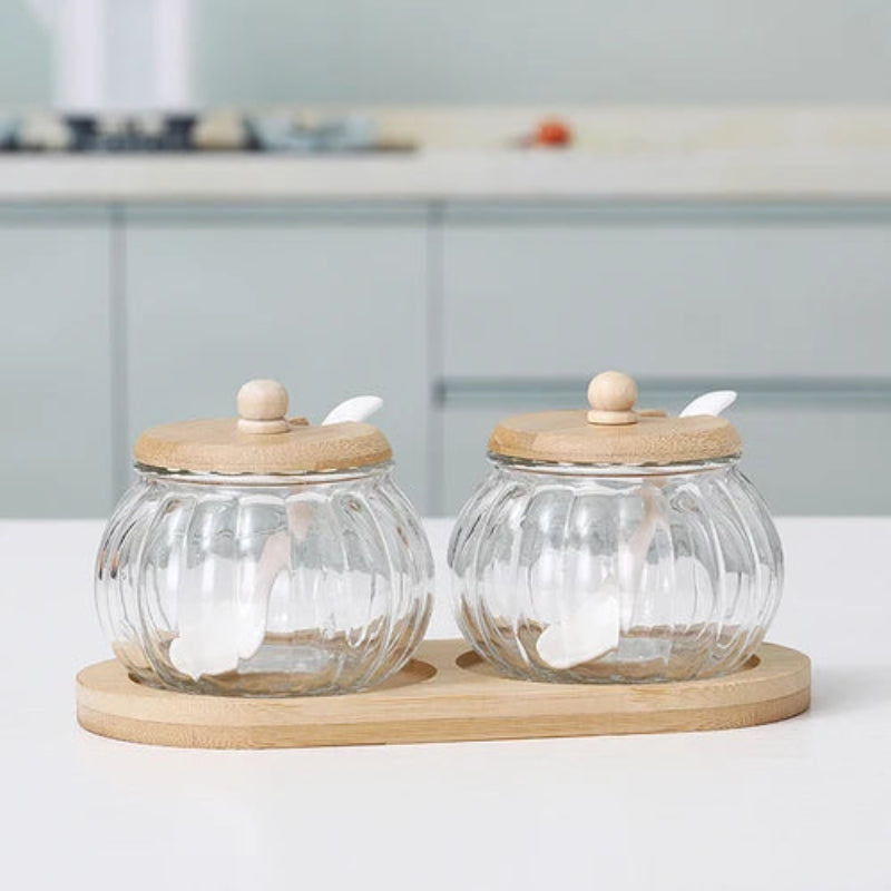 Two empty pumpkin-shaped glass jars with wooden lids and spoons, on a bamboo tray.