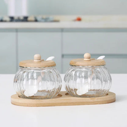 Two empty pumpkin-shaped glass jars with wooden lids and spoons, on a bamboo tray.