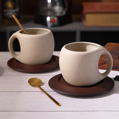 Two ceramic mugs with handles on brown saucers, accompanied by a gold spoon on a wooden surface.
