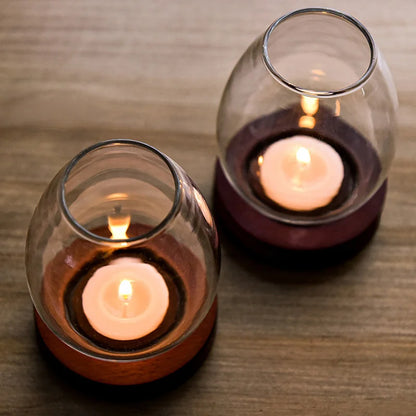 Two glass candle holders with wooden bases, each containing a lit tea light candle, placed on a wooden surface. The warm glow of the candles creates a cozy and intimate atmosphere.