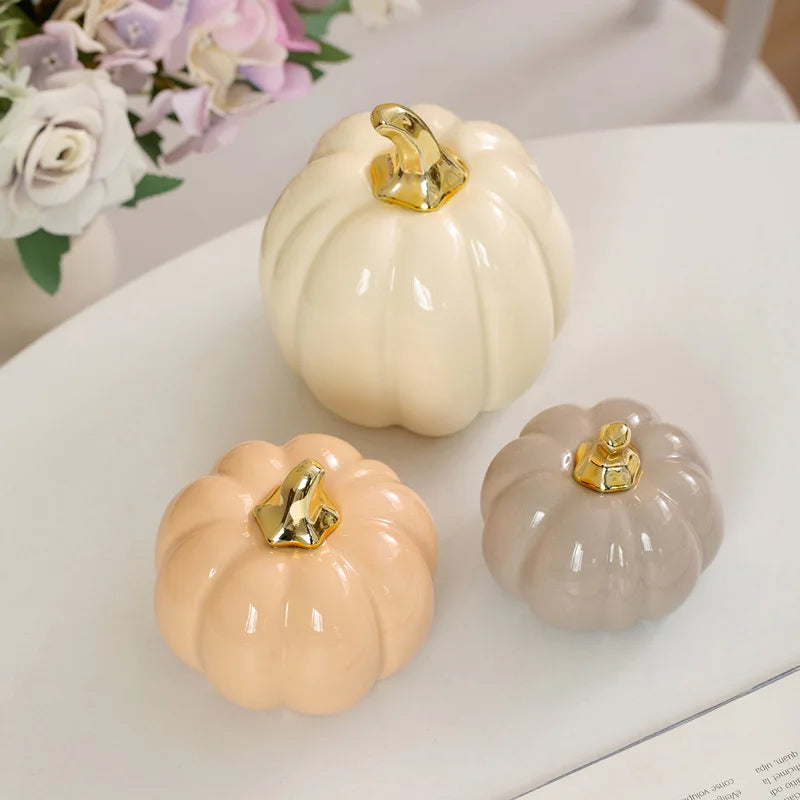Three ceramic pumpkins, colored cream, peach, and gray, with gold stems, are arranged on a white surface, with a blurred floral arrangement in the background.