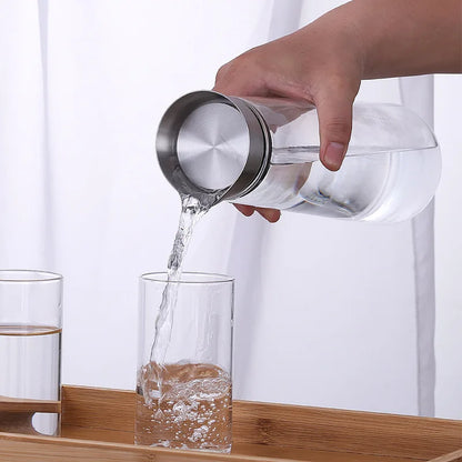 A hand pouring water from a glass carafe with a metal lid into a tall glass.