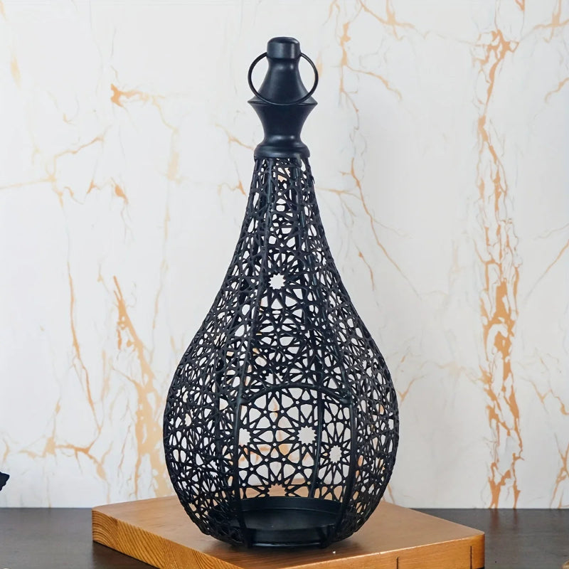 A black, intricately designed metal lantern with a decorative base sits on a wooden surface against a light, subtly patterned wall.
