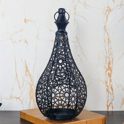 A black, intricately designed metal lantern with a decorative base sits on a wooden surface against a light, subtly patterned wall.