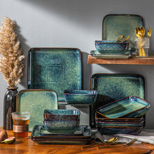 A collection of green and blue square and rectangular dinnerware, with gold cutlery, is displayed on a wooden surface and a shelf.