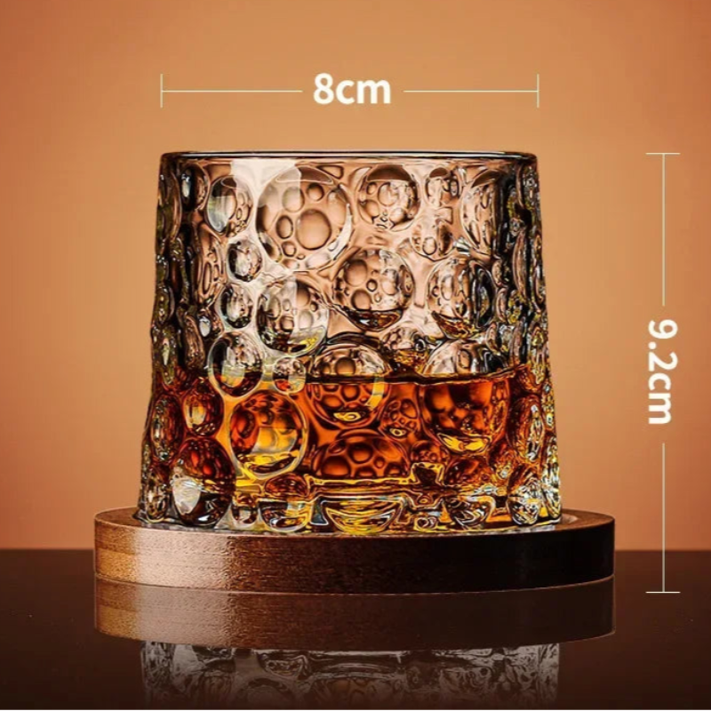 A whiskey glass with a bubble-like texture and amber liquid rests on a wooden coaster, with its dimensions noted.