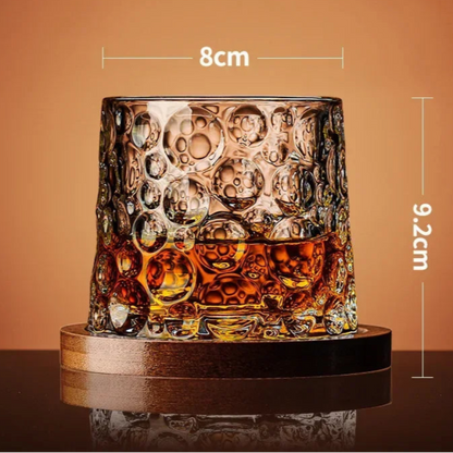 A whiskey glass with a bubble-like texture and amber liquid rests on a wooden coaster, with its dimensions noted.