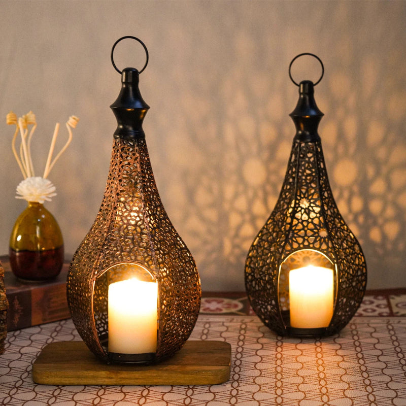 Two intricate metal lanterns with lit candles cast ornate shadows, with one lantern on a wooden base and a decorative vase in the background.