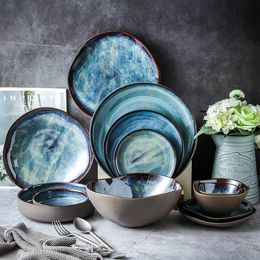 Set of blue ceramic dinnerware on a gray surface with a vase of flowers and silverware.