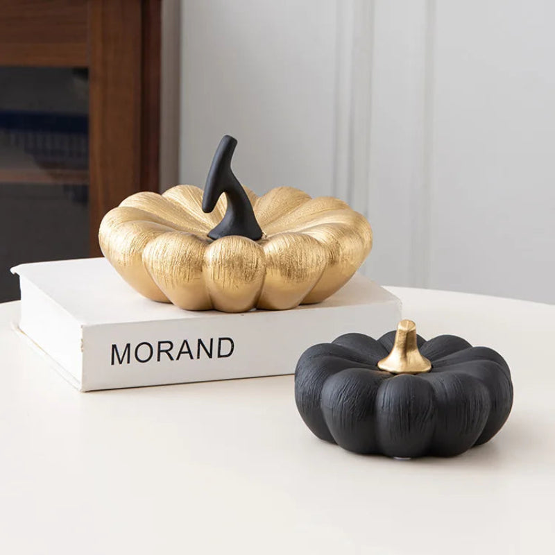 A black pumpkin with a gold stem sits on a table in front of a gold pumpkin with a black stem, which is placed on a book.