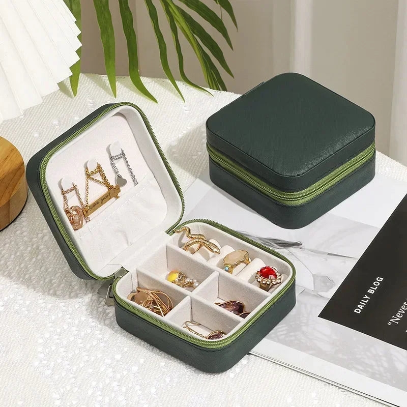 Two dark green, square, zippered jewelry boxes, one open to show organized compartments with various rings and earrings.