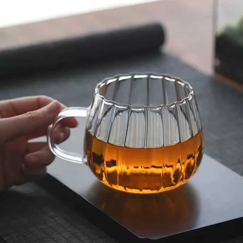 A hand holding a clear, ribbed glass mug with a clear handle, filled with a golden liquid, on a black tray.
