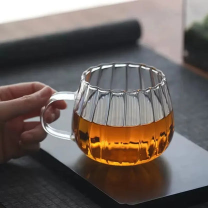 A hand holding a clear, ribbed glass mug with a clear handle, filled with a golden liquid, on a black tray.