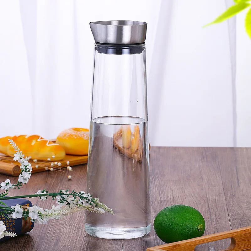 A glass carafe with a metal lid, filled with water, on a wooden table with baked goods and a lime.