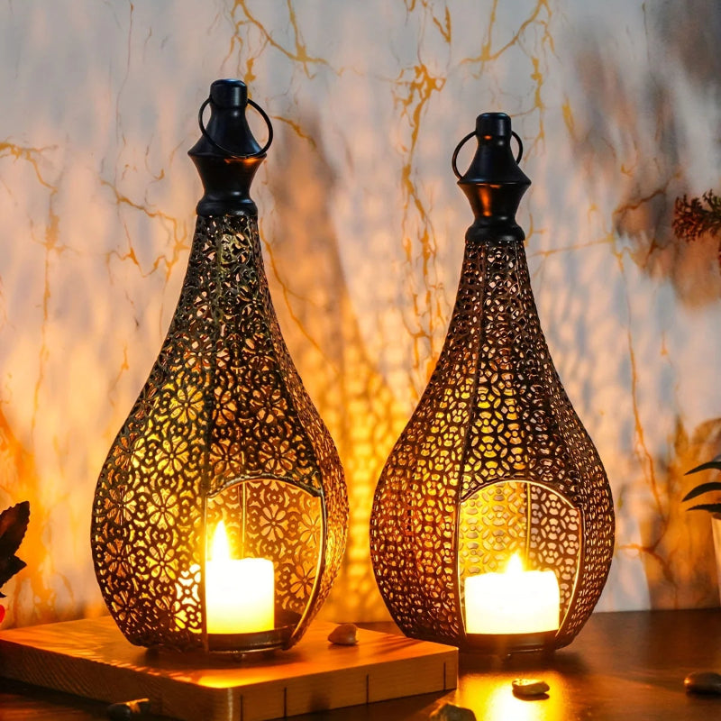 Two ornately designed black metal lanterns, each containing a lit candle, sit on a wooden surface against a light-colored, subtly patterned background.