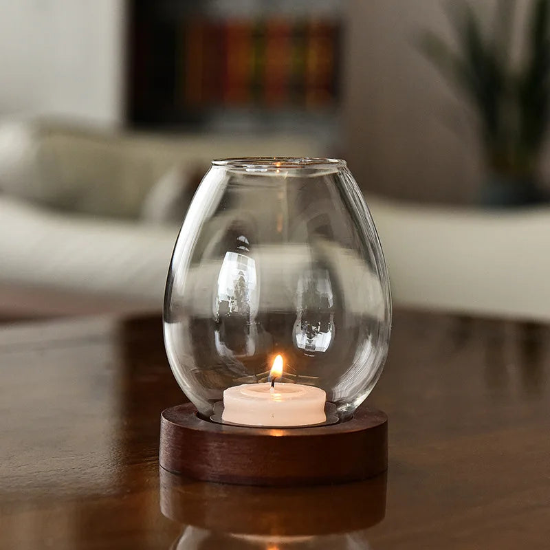 A modern glass candle holder with a wooden base, featuring a softly glowing tealight candle. The transparent glass reflects the surrounding cozy living space, adding a warm and inviting ambiance.