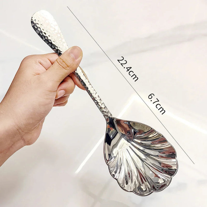 A hand holding an ornate, hammered silver serving spoon with a scalloped, shell-shaped head. The image is labeled with its dimensions.