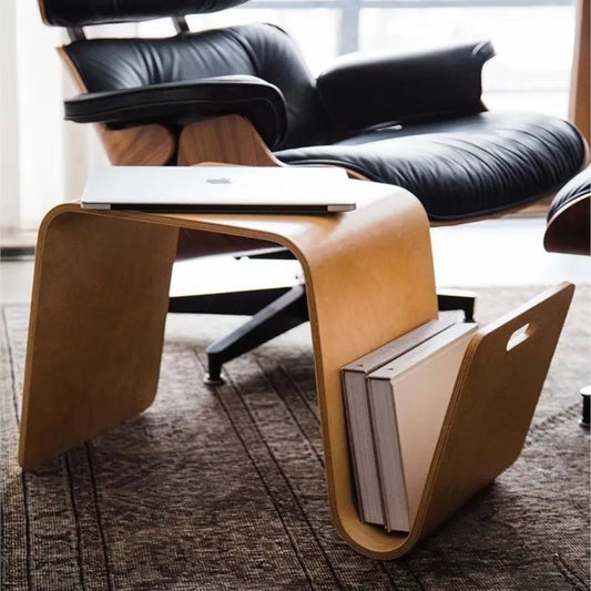 A light wood, bent plywood side table with a laptop on top and books stored in its curved base, next to a black leather lounge chair.