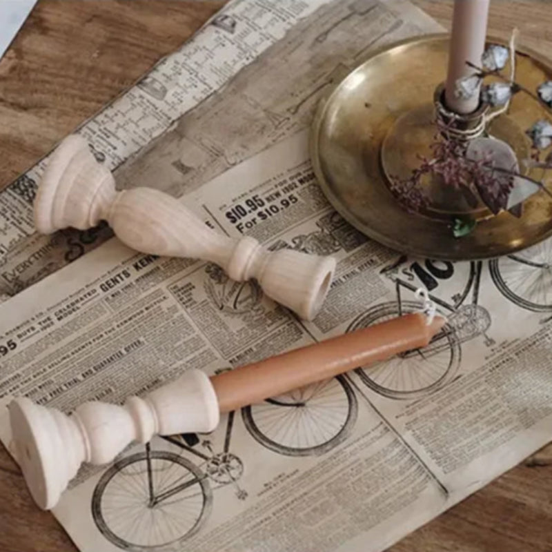 A flat lay shows two turned wooden candle holders next to a brass candle holder with a lit pink taper candle. The objects rest on vintage-style paper with bicycle illustrations and text.