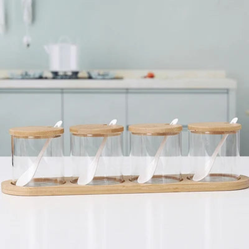 Four empty cylindrical glass jars with wooden lids and spoons, on a bamboo tray.
