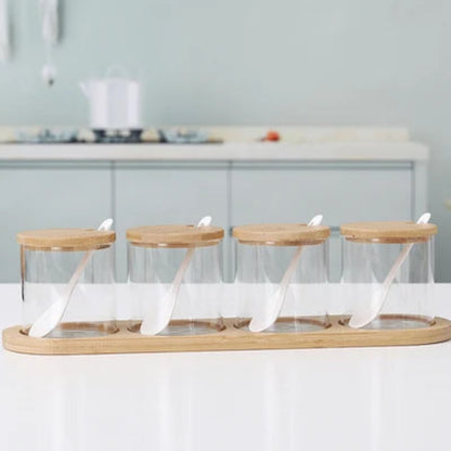 Four empty cylindrical glass jars with wooden lids and spoons, on a bamboo tray.