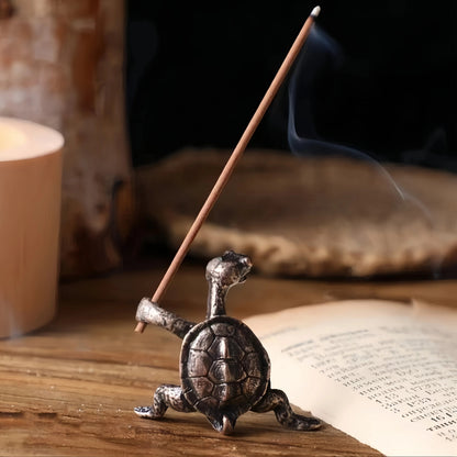 A lit incense stick held by a bronze-colored turtle figurine, with smoke curling upward, on a wooden surface with a book.