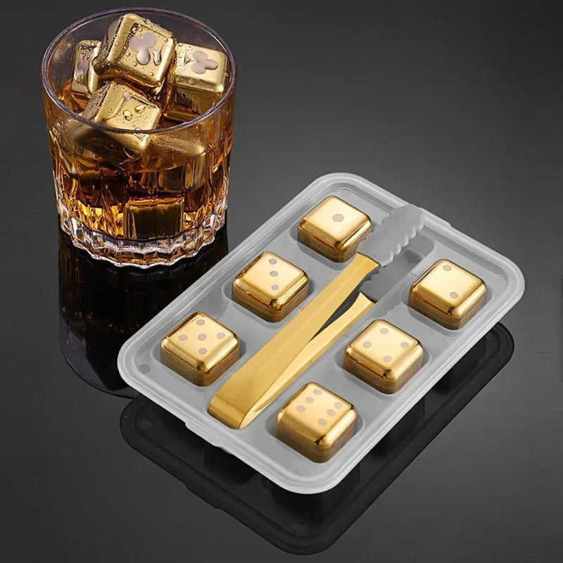 Gold ice cubes in a glass with a tray containing more gold ice cubes and a gold tong on a dark background.