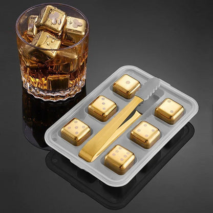 Gold ice cubes in a glass with a tray containing more gold ice cubes and a gold tong on a dark background.