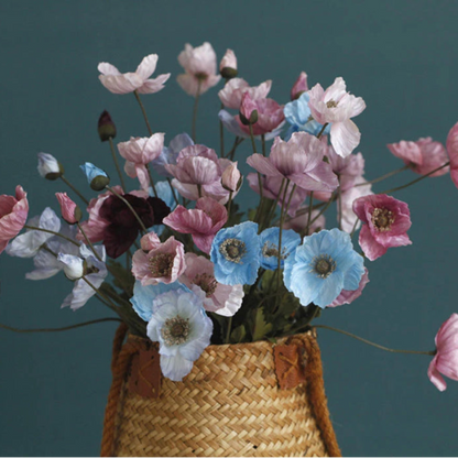 A wicker basket holds a bouquet of various colored poppy flowers, including shades of pink, purple, and blue, against a dark teal background.