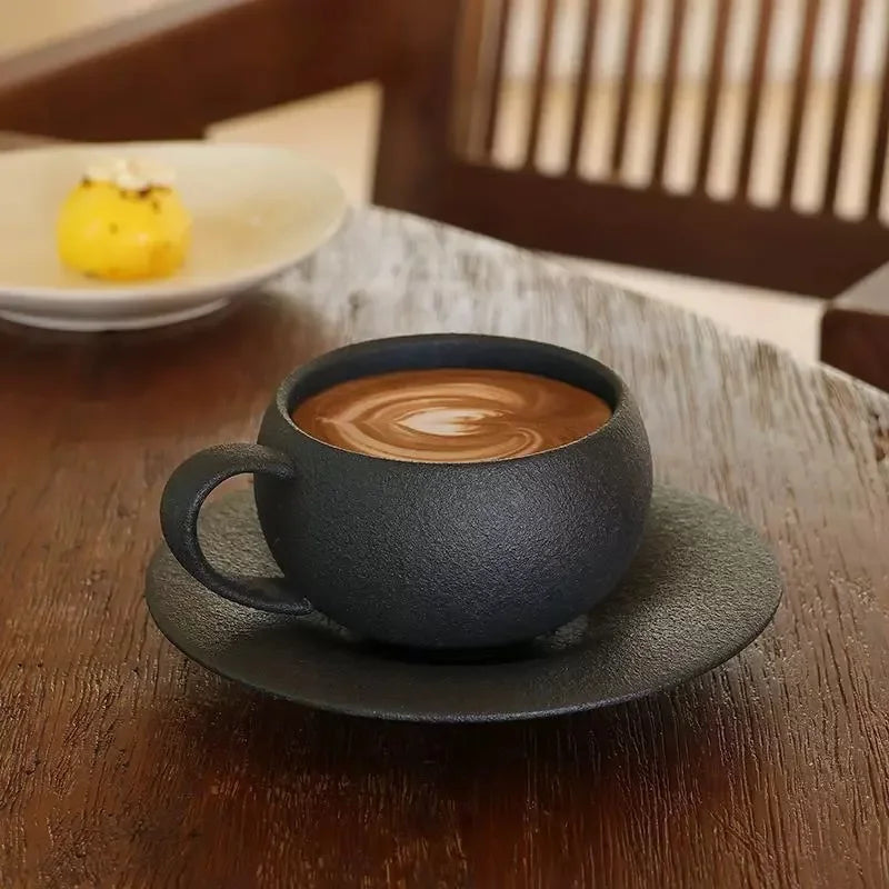 A black textured coffee cup with latte art on a matching saucer, next to a small yellow pastry.