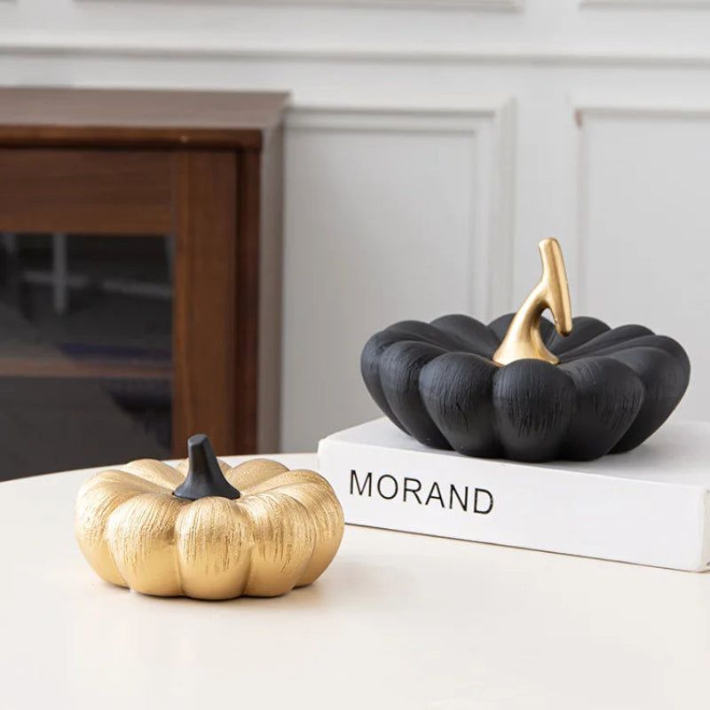 A textured gold pumpkin with a black stem sits on a table next to a black pumpkin with a gold stem, which is placed on a book.