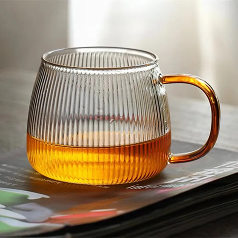 A single ribbed glass mug with an amber handle, containing a golden-colored liquid, resting on top of a book or magazine.