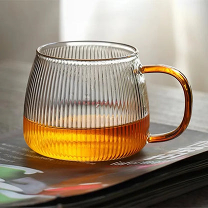 A single ribbed glass mug with an amber handle, containing a golden-colored liquid, resting on top of a book or magazine.