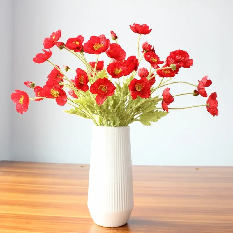 A white ribbed vase holds a bouquet of red poppies on a wooden surface.
