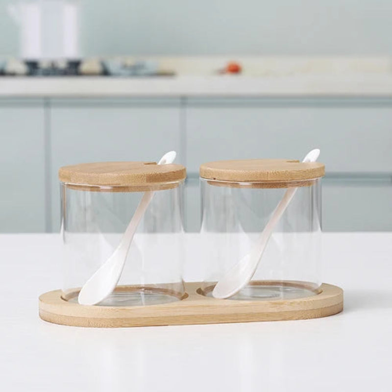 Two empty cylindrical glass jars with wooden lids and spoons, on a bamboo tray.